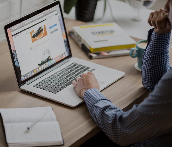 Persoon die een laptop gebruikt voor online leren, met notitieblok en koffie op een houten bureau, een moderne werkplek.