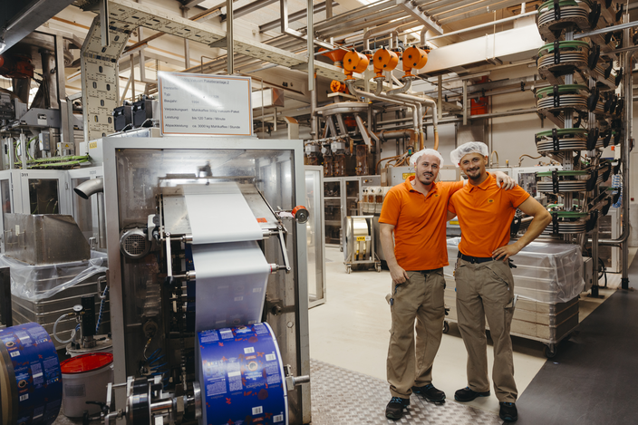 Twee fabrieksarbeiders in oranje shirts poseren trots in een moderne verpakkingsfaciliteit met machines op de achtergrond.