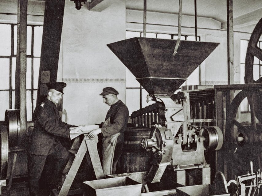 Historická scéna výroby kávy zobrazujúca pracovníkov a stroje v historickej pražiarni. Dedičstvo spoločnosti J.J. Darboven.