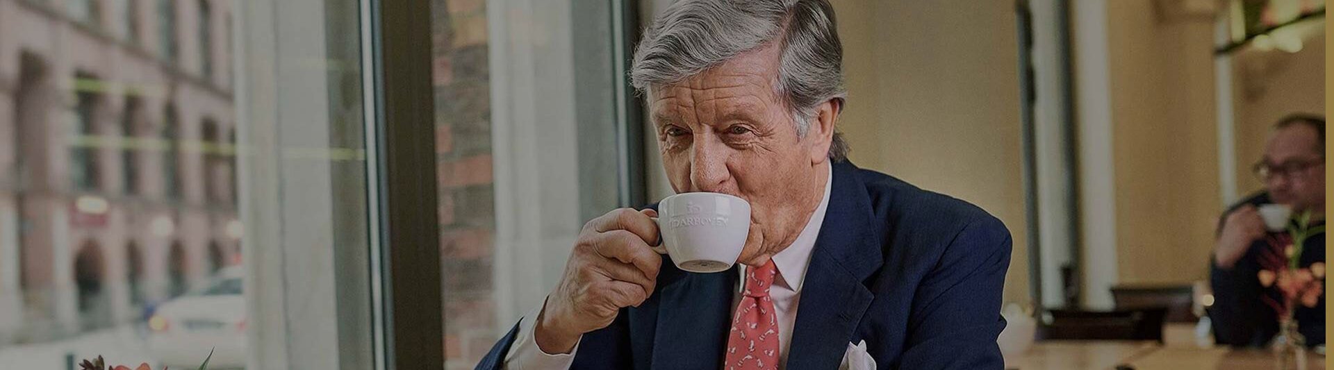 Wyrafinowany mężczyzna delektujący się kawą w kawiarni, otoczony eleganckim wystrojem i widokiem na ulicę miasta.
