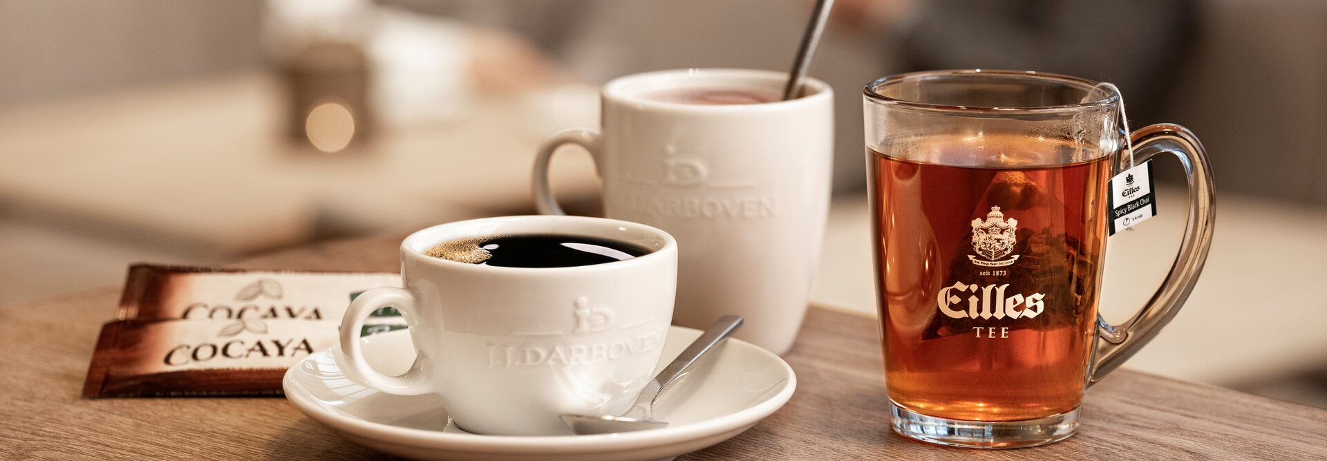 Ein schwarzer Kaffee und ein Glas Tee stehen auf einem Holztisch, und im verschwommenen Hintergrund unterhalten sich zwei Personen.