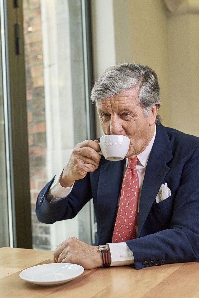 A distinguished man enjoys a cup of coffee at a café, featuring a stylish interior and fresh flowers on the table.