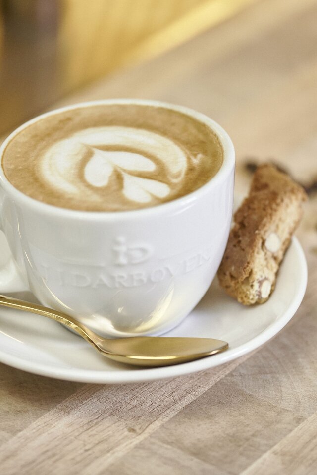 A warm latte with latte art, served in a white cup with a biscuit and coffee beans on a wooden table.