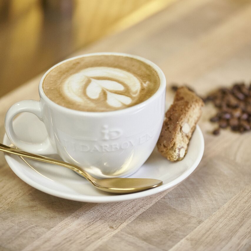 Ein warmer Milchkaffee mit Latte Art, serviert in einer weißen Tasse mit einem Keks und Kaffeebohnen auf einem Holztisch.