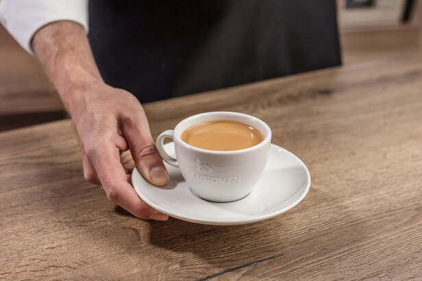 Eine Hand serviert eine Tasse Kaffee auf einer Untertasse, die auf einem Holztisch steht und die satte Farbe des Gebräus zur Geltung bringt.