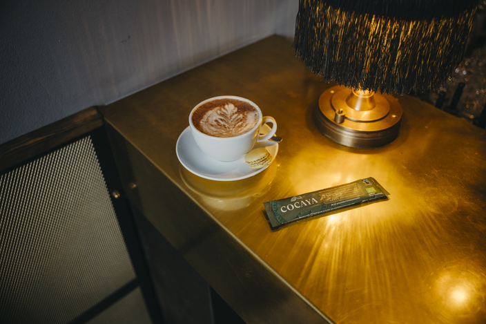 Delicious latte with latte art beside a macaron and a Cocaya health snack bar on a stylish gold table.