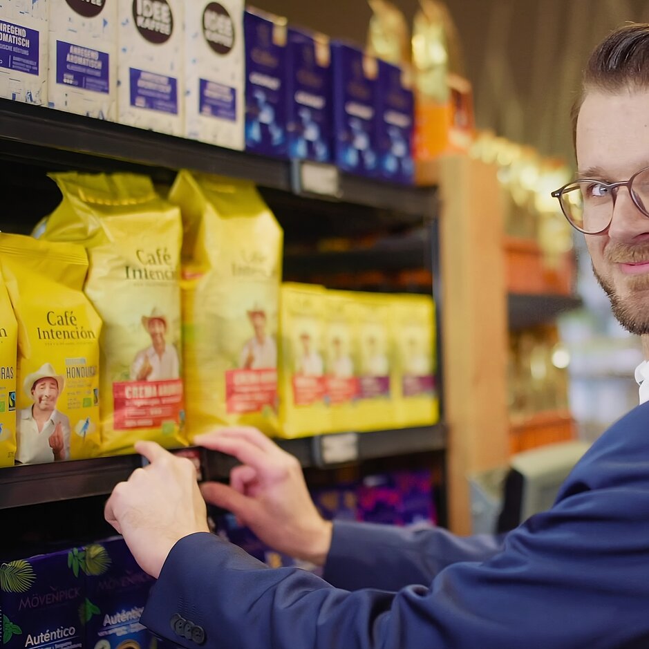 Man in a suit selecting coffee from a shelf, showcasing a variety of colourful coffee brands in a store.