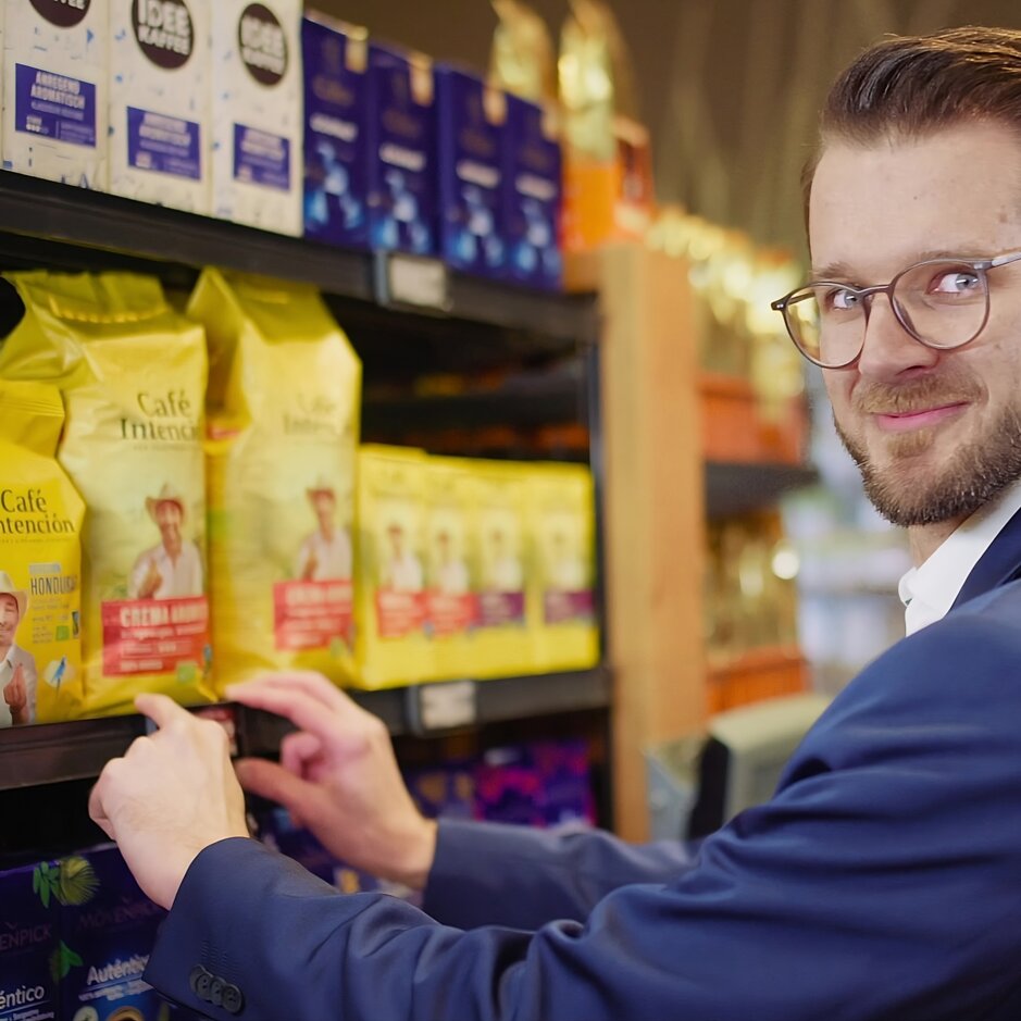 Man in a suit selecting coffee from a shelf, showcasing a variety of colourful coffee brands in a store.