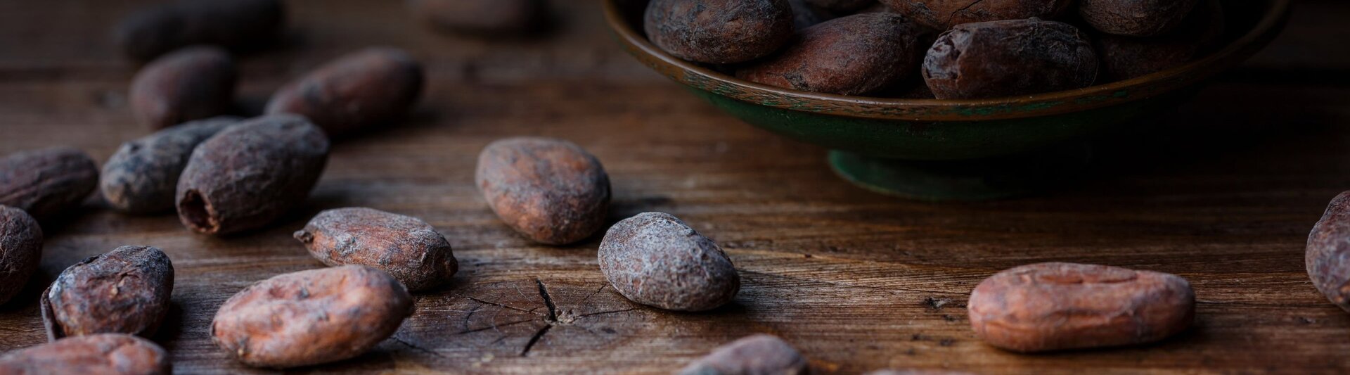 Raw cocoa beans scattered on a rustic wooden surface, with a bowl filled with more cocoa beans in the background.
