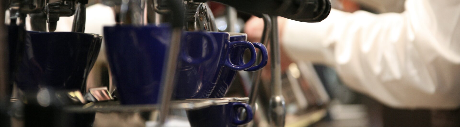Barista bereidt espresso met blauwe kopjes op een espressomachine in een druk café.