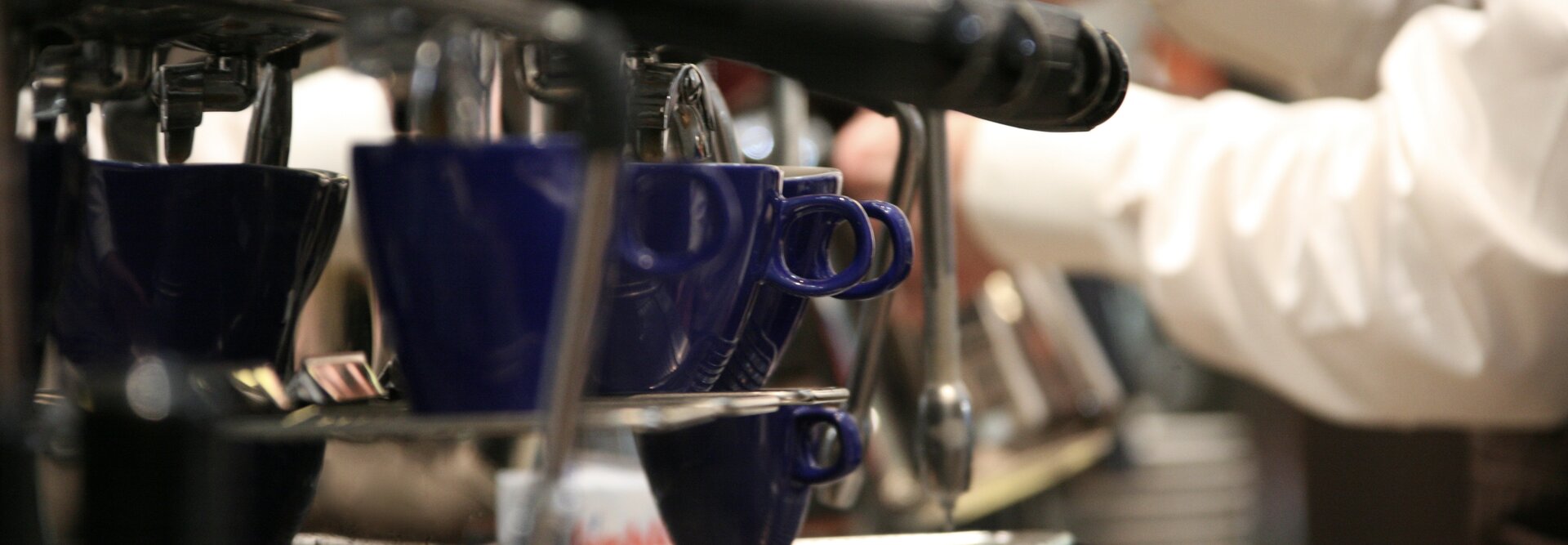 Barista bei der Zubereitung von Espresso mit blauen Tassen an einer Espressomaschine in einem belebten Café.