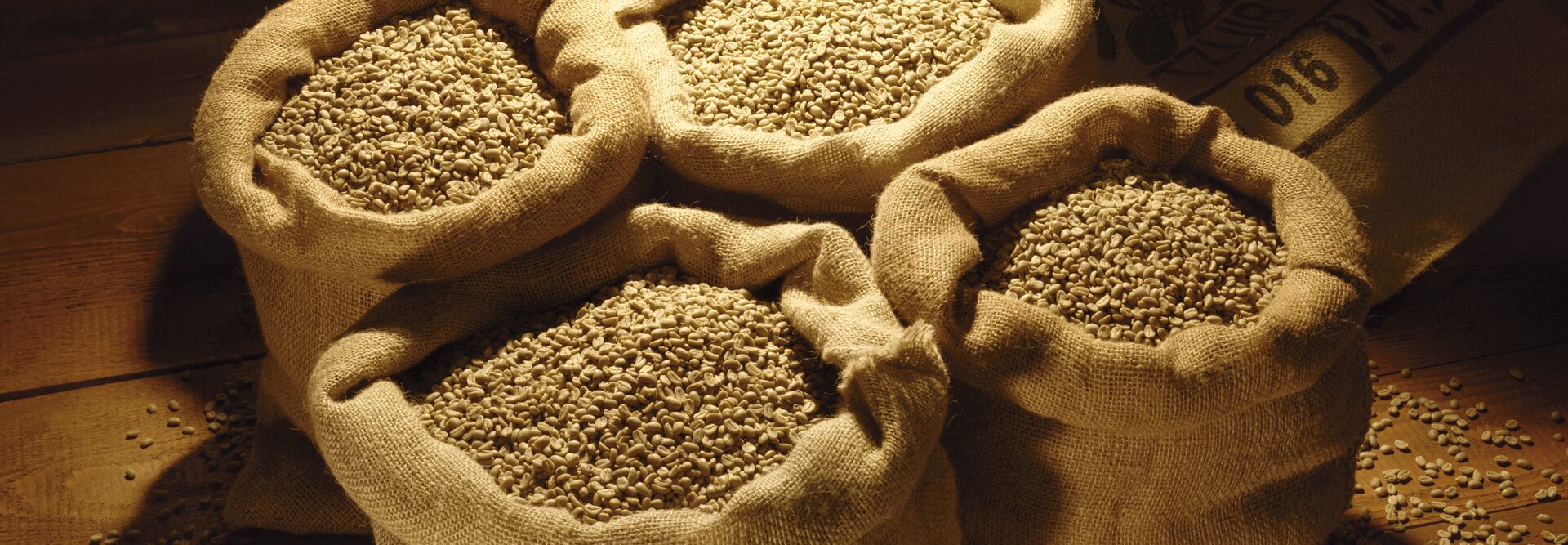 Mit rohen Kaffeebohnen gefüllte Säcke, die auf einer Holzfläche angeordnet sind und von einer dramatischen Beleuchtung unterstrichen werden. Ideal für Kaffee-Liebhaber.