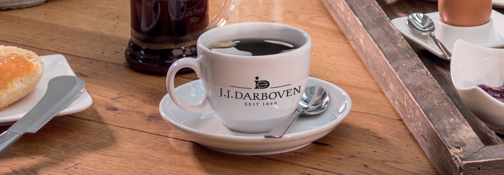 J.J. Darboven-Kaffeetasse auf einem Holztisch, daneben ein Frühstücksbrot, Käse und ein Ei.