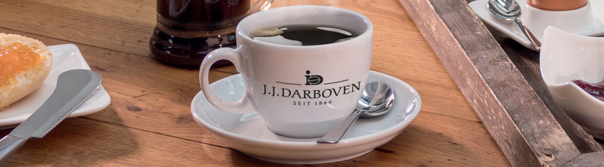 J.J. Darboven coffee cup on a wooden table, alongside a breakfast spread featuring bread, cheese, and an egg.