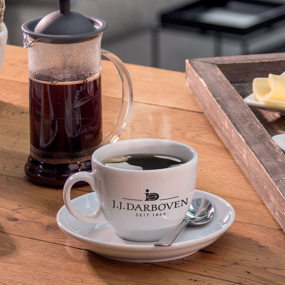 J.J. Darboven-Kaffeetasse auf einem Holztisch, daneben ein Frühstücksbrot, Käse und ein Ei.