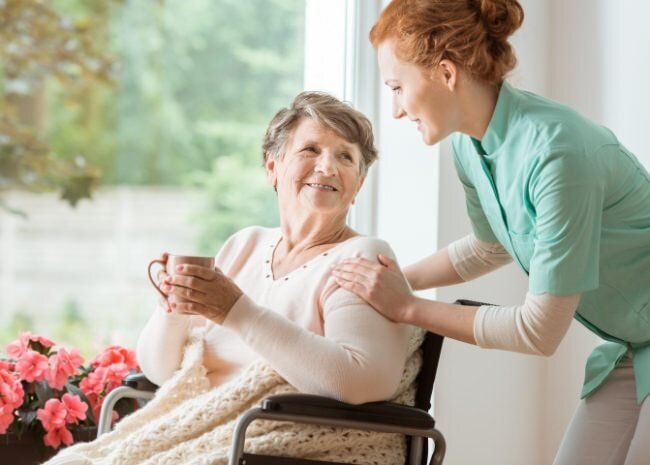A caregiver smiles and supports a senior woman holding a cup in a bright, cheerful room with flowers.