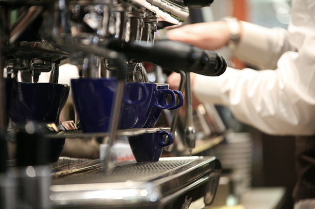 Barista, der mit einer professionellen Espressomaschine und blauen Tassen in einer belebten Café-Atmosphäre Kaffee zubereitet.
