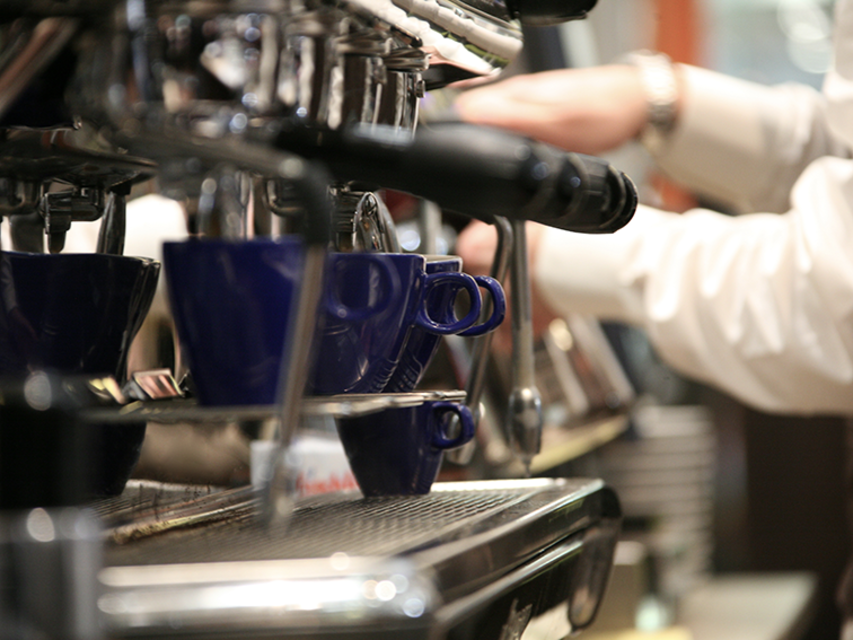 Barista, der mit einer professionellen Espressomaschine und blauen Tassen in einer belebten Café-Atmosphäre Kaffee zubereitet.