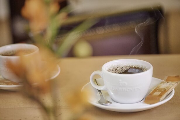 Dampfender Kaffee in einer weißen Tasse auf einem rustikalen Tisch, ergänzt durch ein Süßstoffpaket und einen unscharfen floralen Hintergrund.