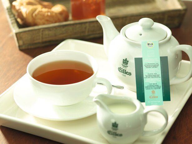 Eine dampfende Tasse mit englischem Bio-Frühstückstee, serviert mit Milch, einer Teekanne und einem Croissant auf einem Holztisch.