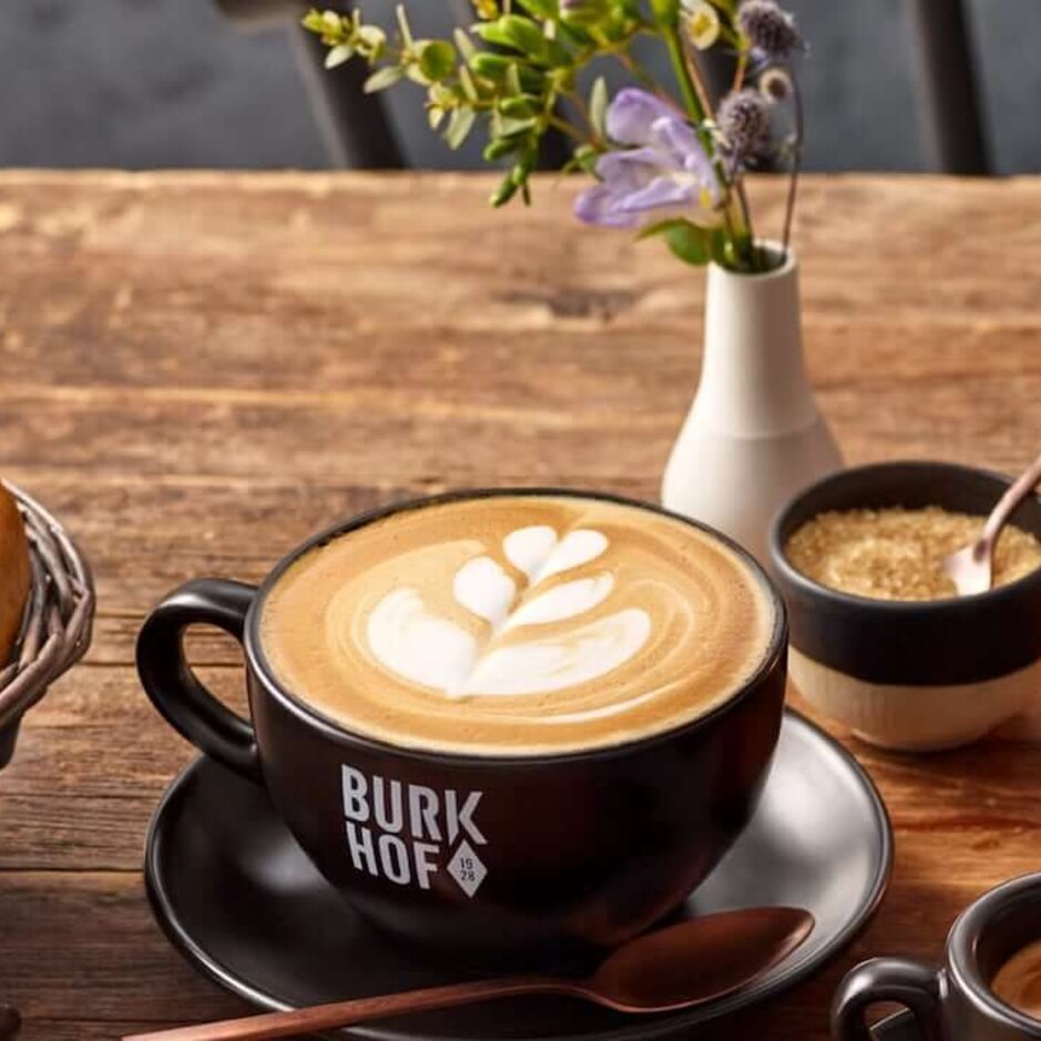 Handwerklich hergestellter Kaffee mit Latte Art, frische Bagels und zarte Blumen auf einem rustikalen Holztisch im Café Burkhof.