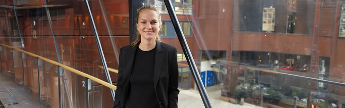 Obchodní profesionál v elegantním černém saku, který sebevědomě stojí v moderní kanceláři se skleněnou architekturou.