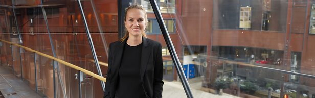 Obchodní profesionál v elegantním černém saku, který sebevědomě stojí v moderní kanceláři se skleněnou architekturou.