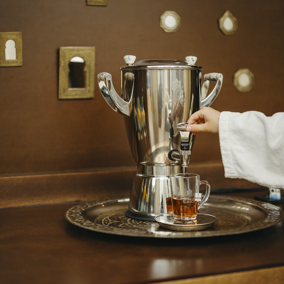 Ręka nalewa herbatę z eleganckiego srebrnego dozownika do szklanej filiżanki na ozdobnej tacy, a obok stoi woda.