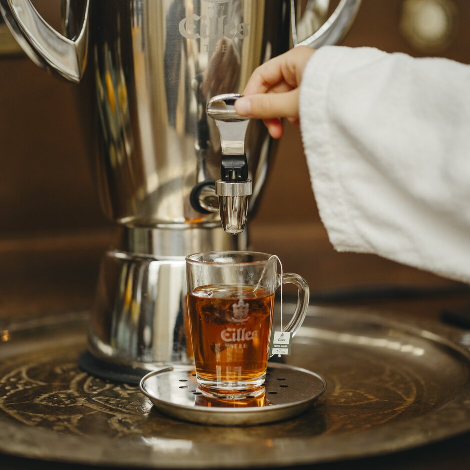Eine Hand gießt Tee aus einem stilvollen Spender in einen Glasbecher und präsentiert das warme Getränk auf einem dekorativen Tablett.