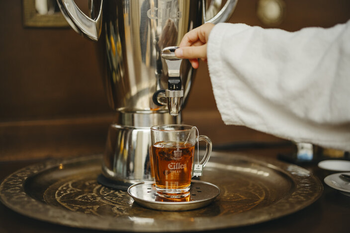 A hand pours tea from a stylish dispenser into a glass mug, showcasing a warm beverage on a decorative tray.