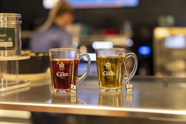 Zwei durchsichtige Tassen mit Cilles-Tee, eine rote und eine grüne, stehen auf einem Tresen in einem Café.