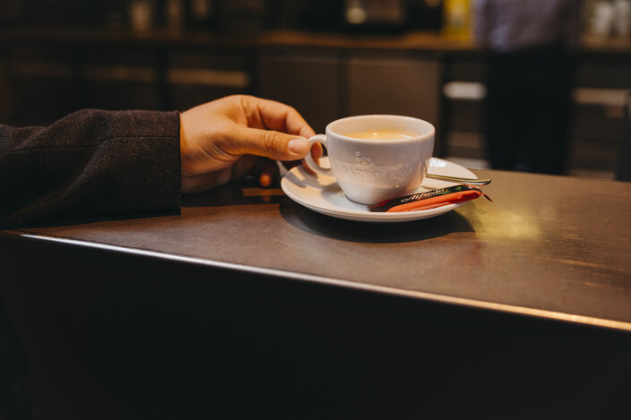 A hand holds a white coffee cup on a cafe countertop, accompanied by a small spoon and sugar stick.