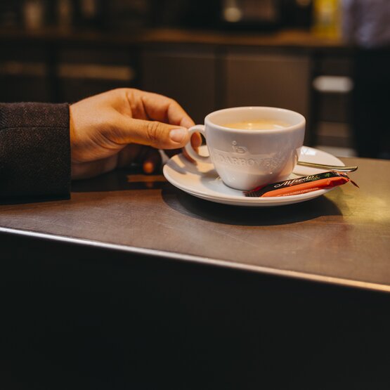 A hand holds a white coffee cup on a cafe countertop, accompanied by a small spoon and sugar stick.
