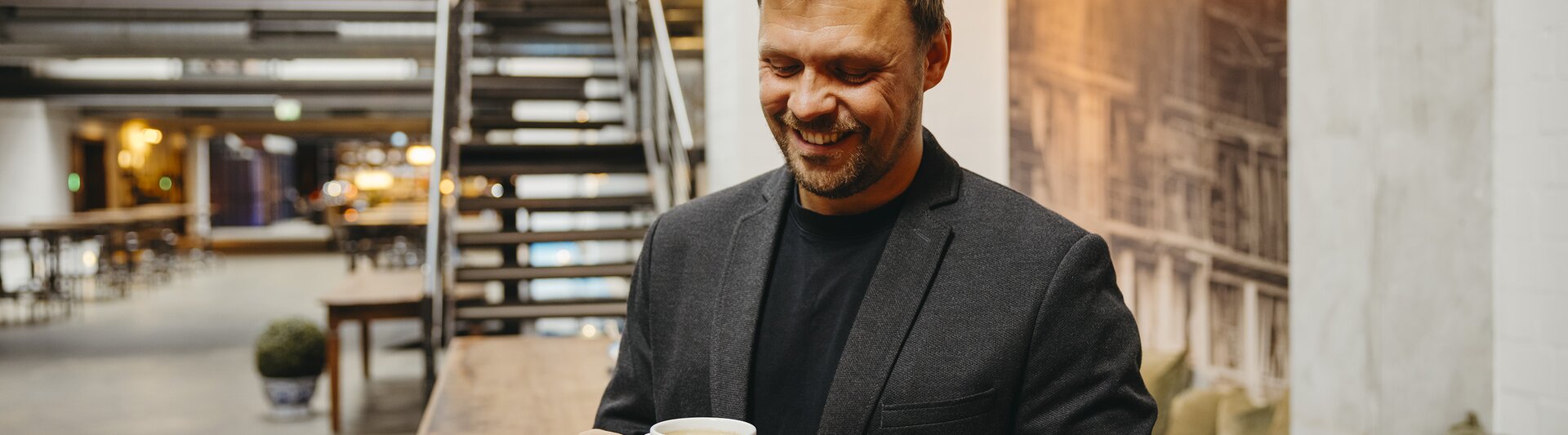 A smiling man in a blazer enjoys a cup of coffee in a stylish, modern café setting with wooden furniture.