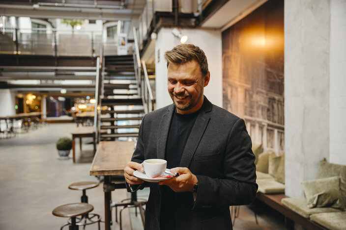 Ein lächelnder Mann in einem Blazer genießt eine Tasse Kaffee in einem stilvollen, modernen Café mit Holzmöbeln.