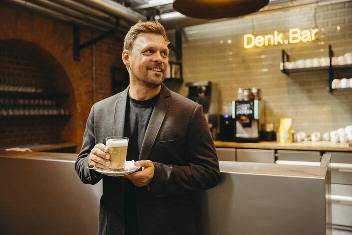 A man in a stylish blazer enjoys a drink at Denk.Bar, showcasing a modern café atmosphere.