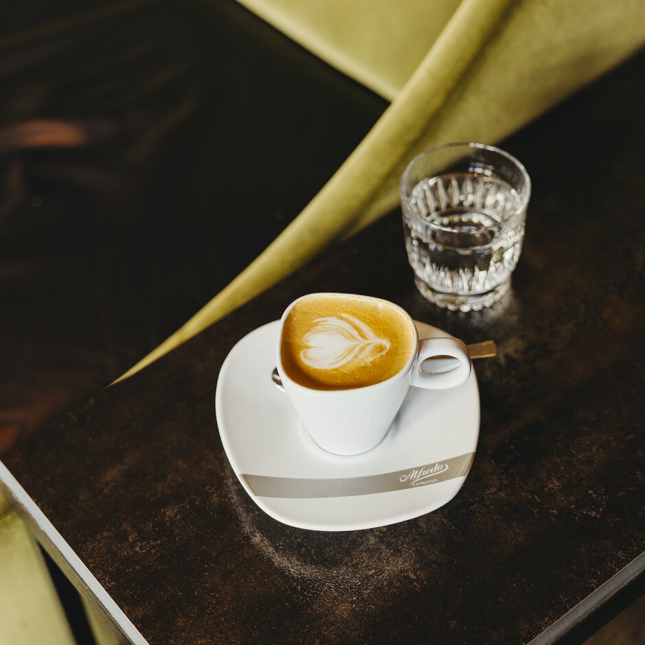 Krásne spracované latte art leží na bielom tanieriku vedľa pohára s vodou na štýlovom drevenom stole.