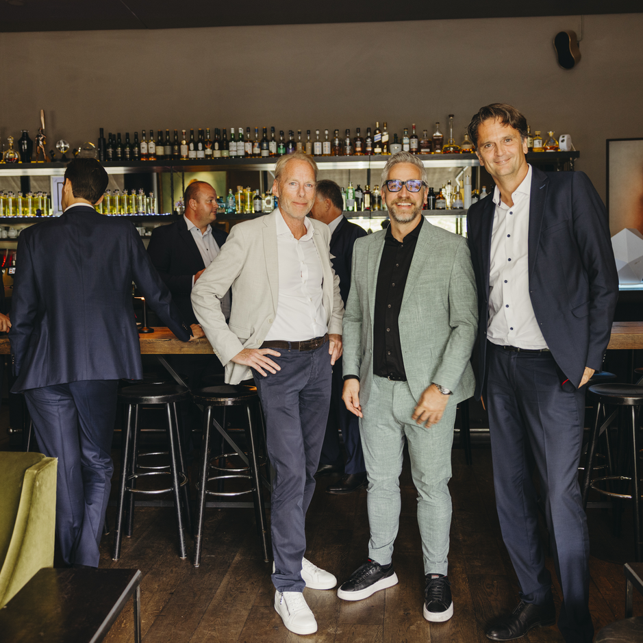 Three men in smart attire pose in a stylish bar, with shelves of bottles in the background and colleagues engaged in conversation.