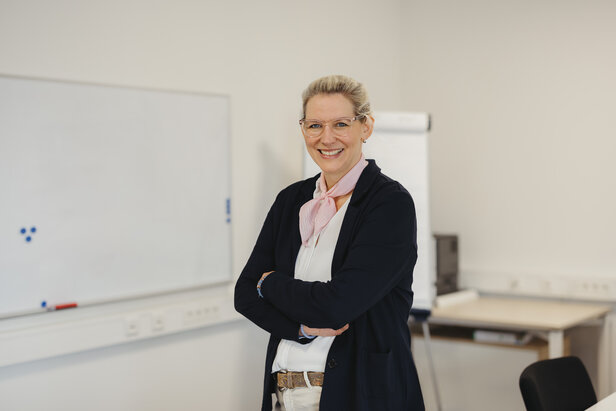 Une professionnelle souriante avec des lunettes pose avec assurance dans un bureau lumineux avec un tableau blanc en arrière-plan.