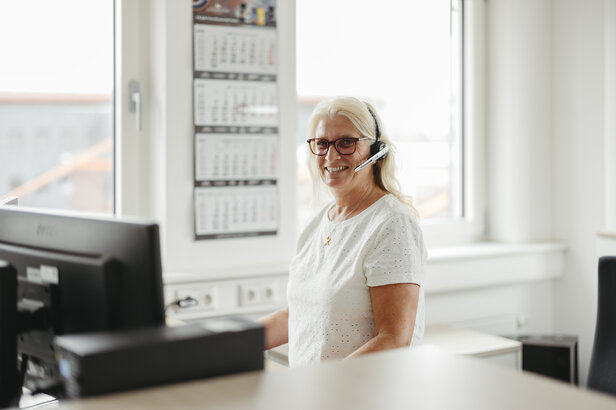 Femme souriante avec des lunettes et un casque à son bureau, travaillant dans un environnement de bureau lumineux.