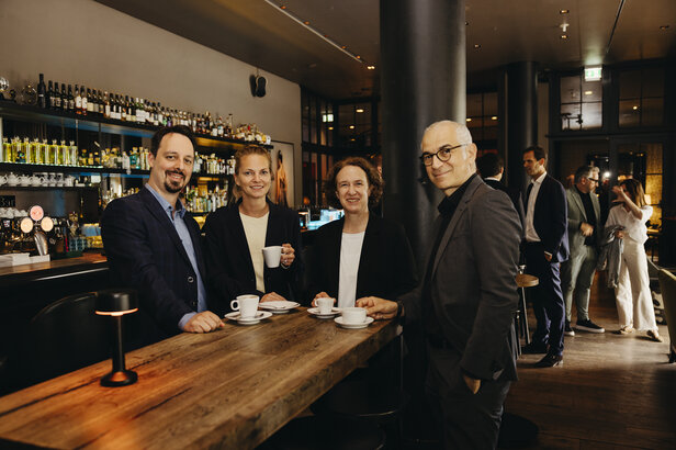 Štyria profesionáli si spoločne vychutnávajú kávu v štýlovom bare, kde vládne živá spoločenská atmosféra.