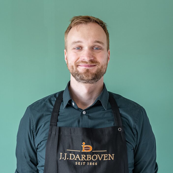 Trainer from J.J. Darboven, smiling against a green background, wearing an apron with the brand logo.