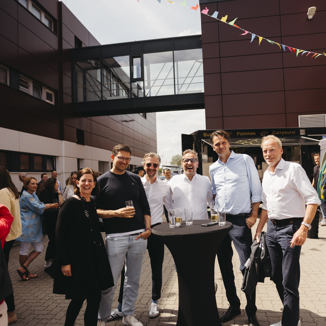 Les invités prennent un verre lors d'un événement en plein air, entourés de banderoles et d'un bâtiment moderne, créant ainsi une atmosphère festive.