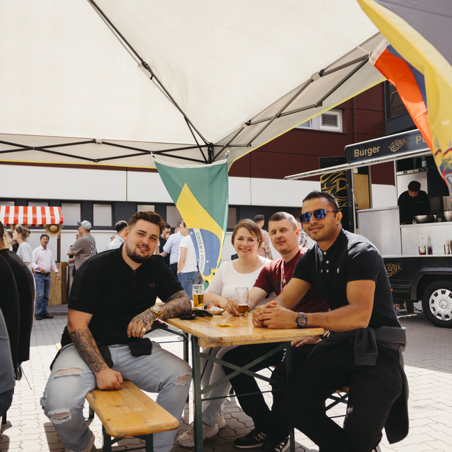 Des amis prennent un verre sur un marché alimentaire animé, sous un auvent, avec un drapeau brésilien en arrière-plan.