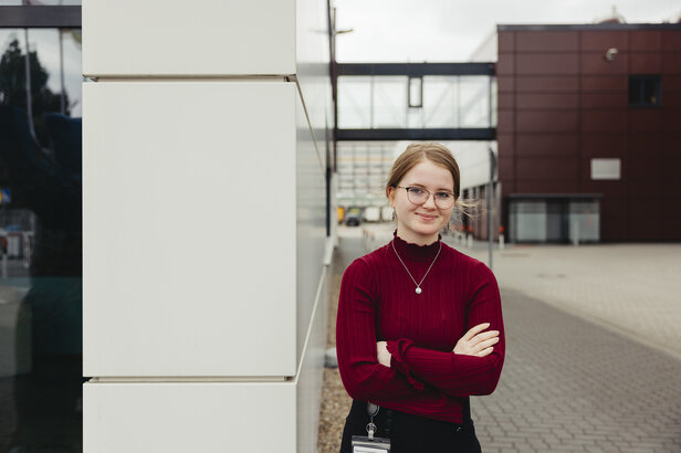 Un jeune professionnel vêtu d'un pull-over rouge se tient avec assurance à l'extérieur, mettant en valeur l'architecture moderne à l'arrière-plan.