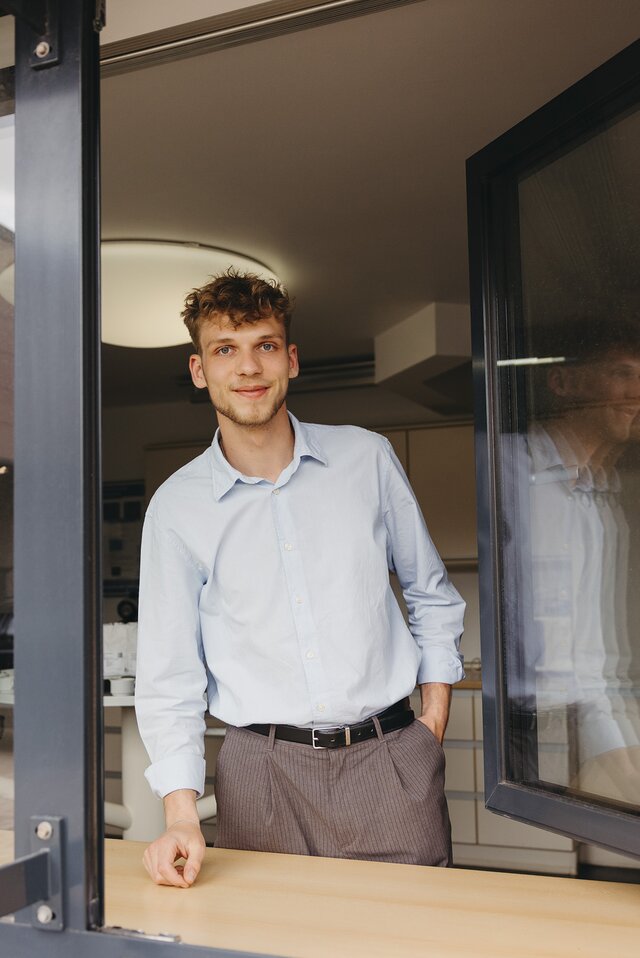 Un jeune homme souriant, vêtu d'une chemise bleu clair, se penche à travers la fenêtre d'un café à l'atmosphère accueillante.