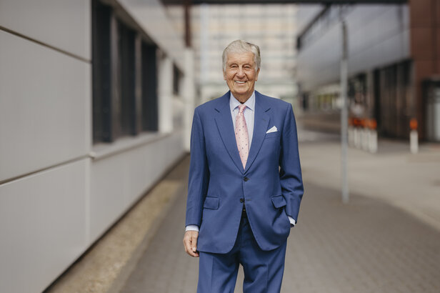 Ein älterer Mann in blauem Anzug und rosa Krawatte lächelt selbstbewusst, während er einen modernen städtischen Weg entlanggeht.