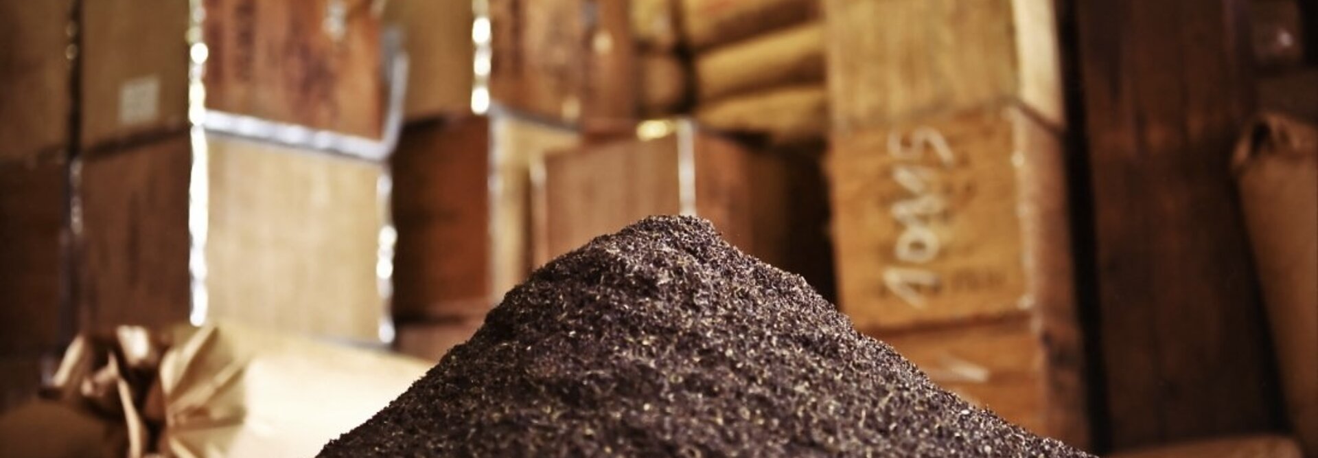 Mountain of dark ground spices in a rustic storage area with stacked boxes in the background.