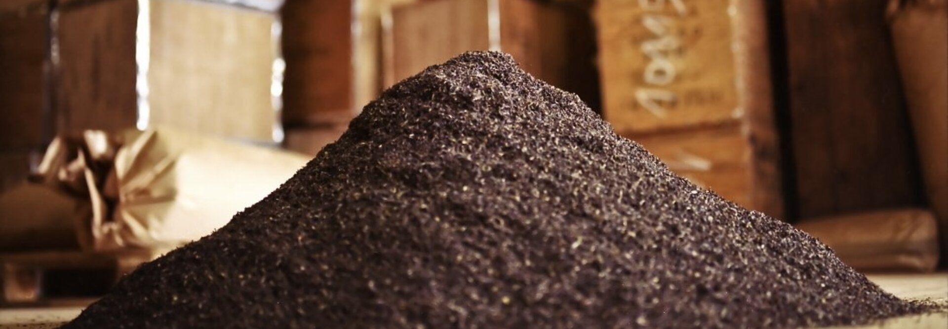 Berg von dunkel gemahlenen Gewürzen in einem rustikalen Lagerraum mit gestapelten Kisten im Hintergrund.