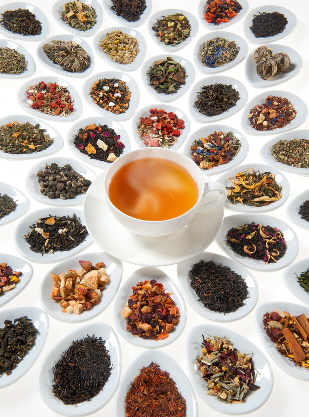 Rôzne sypané čaje a bylinky naaranžované okolo šálky čaju v pare na elegantných bielych podšálkach.
